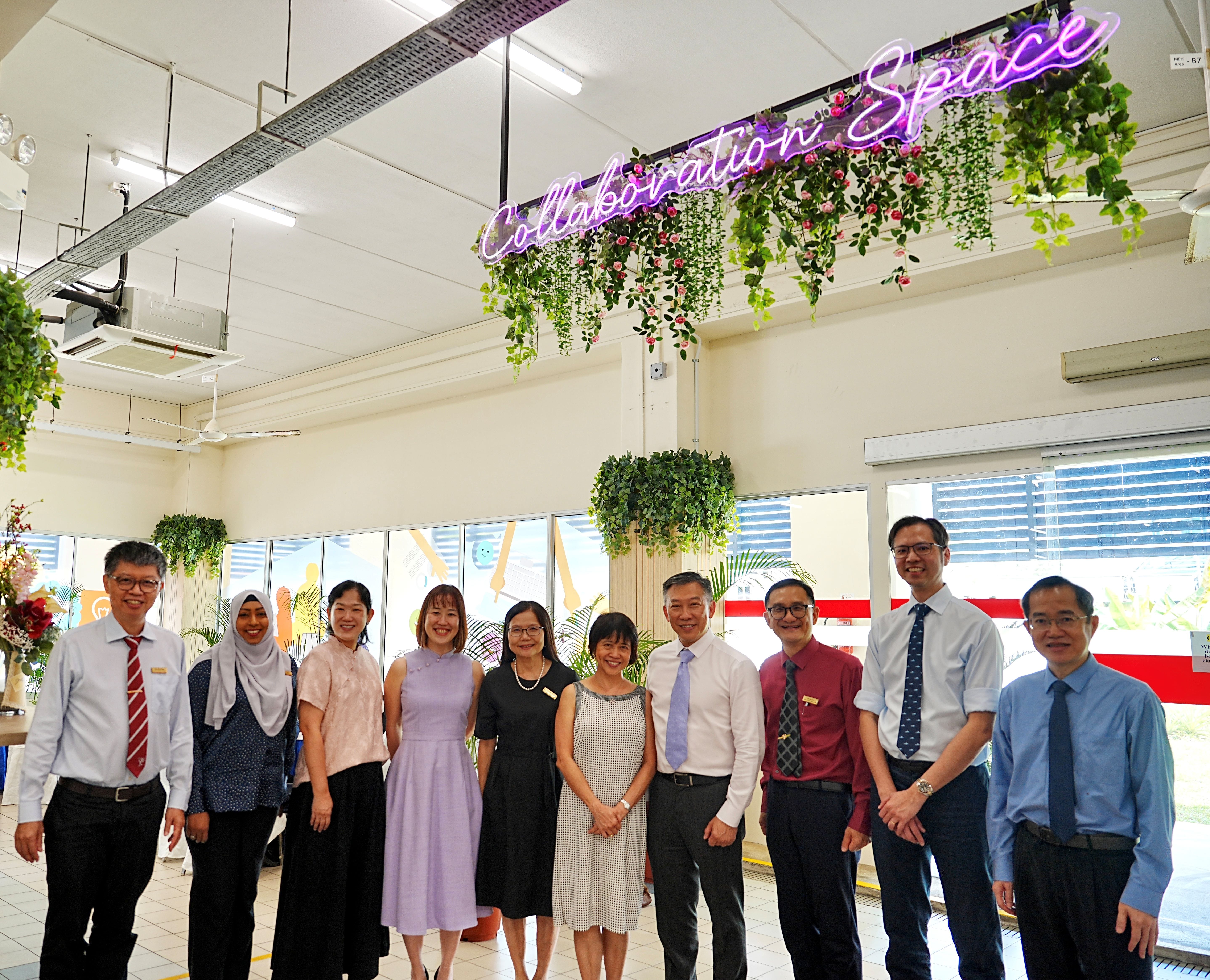 A group of ten people — a mix of men and women in formal attire — pose together indoors beneath a glowing purple neon sign reading "Collaboration Space," decorated with hanging artificial greenery and flowers.
